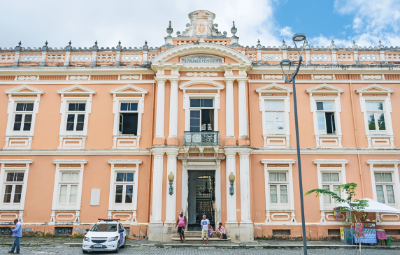 Imagem: Fotografia. Fachada de um prédio de piso duplo, parte superior com arabescos, grandes janelas verticalizadas envidraçadas e entrada com abertura espaçosa à frente de uma pequena escada que dá acesso à calçada, onde estão algumas pessoas. No estacionamento, há um carro. Do lado direito, uma pequena árvore e uma barraca de vendedor ambulante.  Fim da imagem.