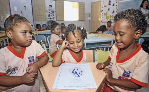 Imagem: Fotografia. Em uma sala de aula com colagens nas paredes, portas e janelas de madeira, três crianças uniformizadas estão sentadas dividindo uma pequena mesa na qual há uma folha com o desenho de um abacaxi. Ao fundo, outras mesas nas quais estão outros alunos. Fim da imagem.