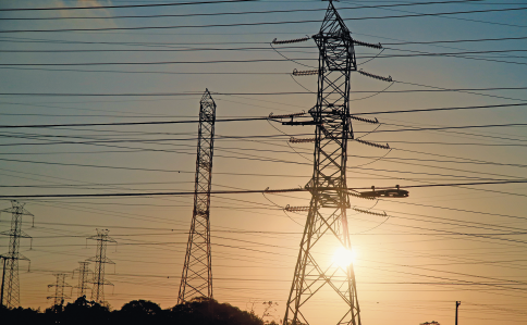 Imagem: Fotografia. Destaque de altas torres de ferro pelas quais passam vários cabos, alguns mais grossos e outros mais finos. No horizonte, o sol.  Fim da imagem.