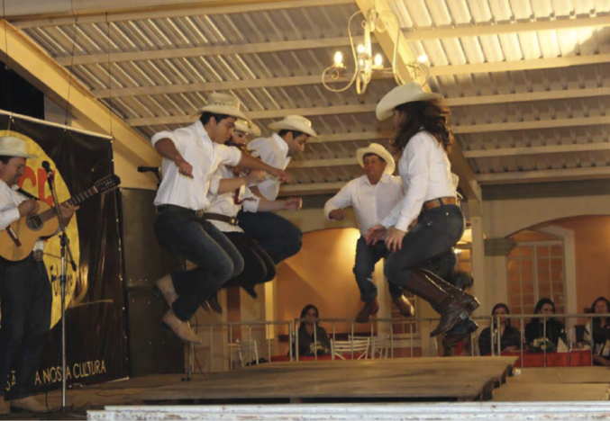 Imagem: Fotografia. Em um palco diante de músicos com violão, um grupo de pessoas, homens e mulheres, dançam sobre um tablado de madeira. Todos usam chapéu com abas curvas, camisa branca, calça escura e sapatos com salto baixo. Ao redor, está a plateia sentada. Fim da imagem.