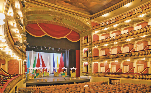 Imagem: Fotografia. Interior de um espaço amplo com pé direito alto e muitas cadeiras vazias diante de um palco em piso superior e decorado. Nas laterais, há quatro andares com espaçosos camarotes. A iluminação do local é amarela e os detalhes decorativos são dourados.  Fim da imagem.