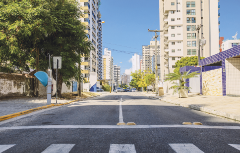 Imagem: Fotografia. Destaque de uma rua pavimenta com sinalização de trânsito. Na calçada há casas, prédios, árvores e telefone público.  Fim da imagem.