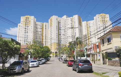 Imagem: Fotografia. Destaque de uma rua pavimentada com muitos carros estacionadas à frente das calçadas. Na região, há muitas casas assobradas e prédios ao fim da rua.  Fim da imagem.