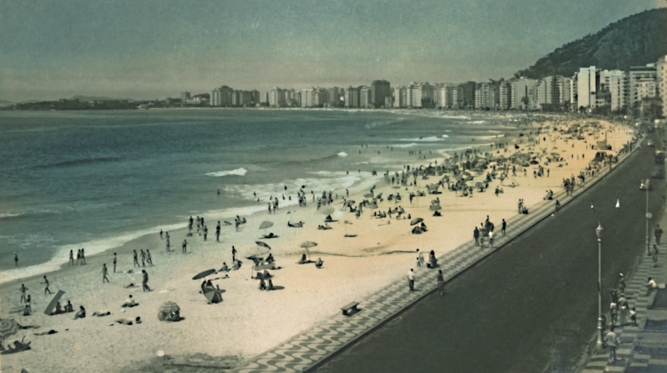 Imagem: Fotografia. Plano aberto da orla de uma praia. A cidade tem muitos prédios e extensa avenida próximo ao calçadão. Muitas pessoas caminham pelo espaço, há guarda-sóis na areia e banhistas na água.  Fim da imagem.