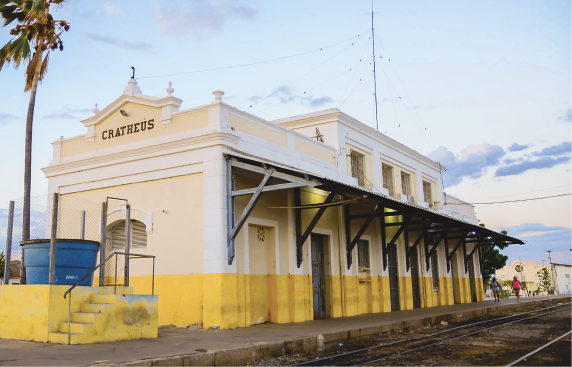 Imagem: Fotografia. Fachada de uma estação de trem. Uma construção comprida com portas e janelas em uma área coberta e com plataforma estreita diante do trilho do trem. Na lateral, há uma pequena escada com acesso a uma porta.  Fim da imagem.