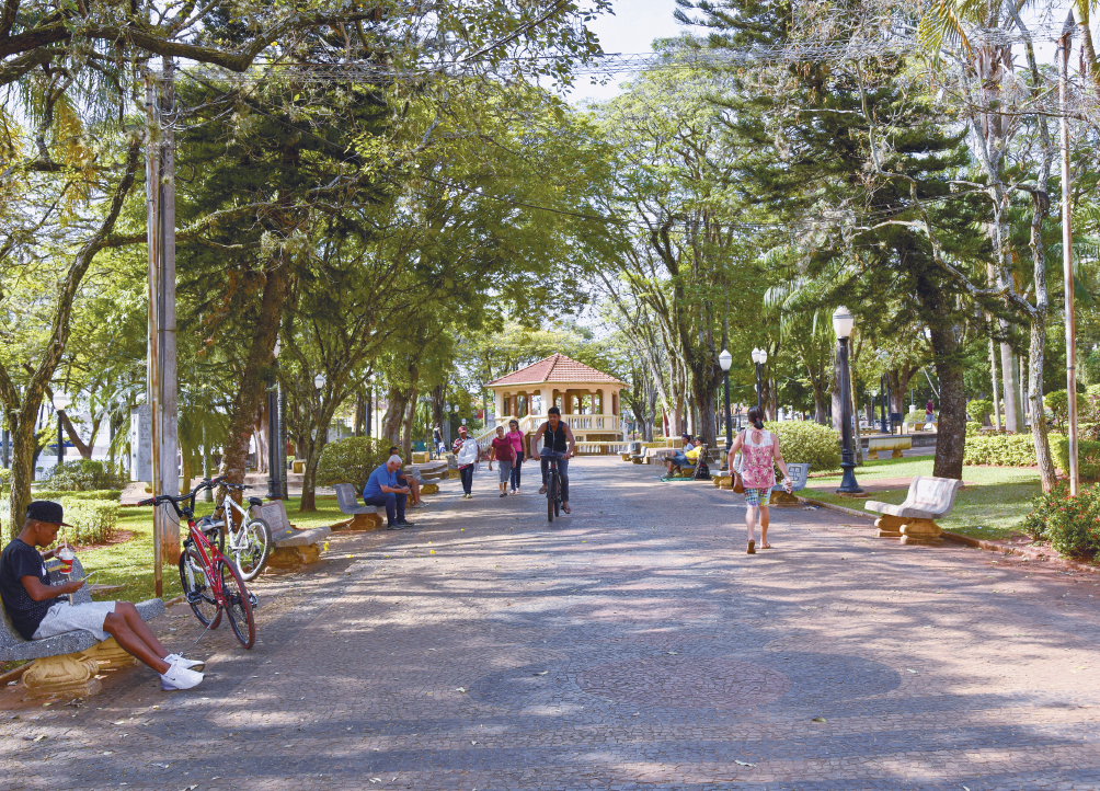 Imagem: Fotografia. Destaque de uma ampla área de passeio por onde transitam algumas pessoas e um ciclista. Nas laterais, bancos, postes de iluminação e muitas árvores espalhadas em uma área gramada. Ao fundo, um coreto. Fim da imagem.