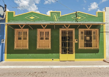 Imagem: Fotografia. Fachada de uma casa térrea com janelas e portas de acesso direto à rua e com grades de proteção.  Fim da imagem.