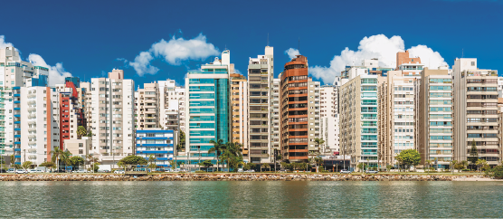 Imagem: Fotografia. Plano aberto da encosta da praia vista do mar. No horizonte, a faixa de areia e muitos prédios altos.  Fim da imagem.