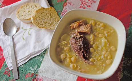 Imagem: Fotografia. Sobre uma toalha estendida nas cores branca, vermelha e verde, está um prato com ensopado e carne e um guardanapo com uma colher e duas fatias de torrada.  Fim da imagem.