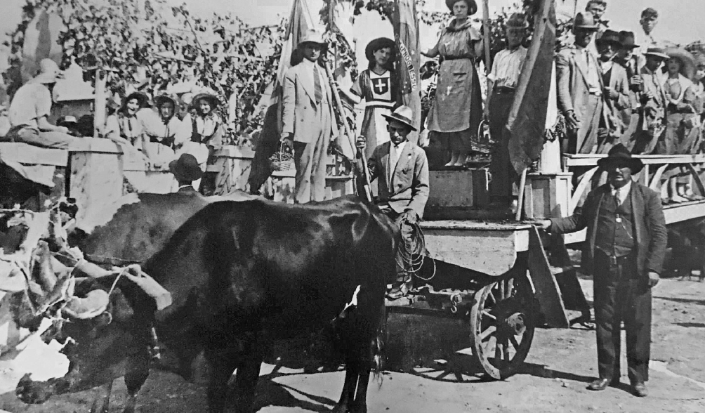 Imagem: Fotografia em preto e branco. Em uma rua, destaque de uma carreta de madeira sobre rodas puxadas por bois. Sobre a estrutura, estão muitas pessoas, homens de terno e chapéu e mulheres de vestido e chapéu, que seguram bandeiras e posam para foto. Ao fundo, algumas mulheres observam. No chão, ao lado da carroceria, um homem de terno e chapéu também olha para a foto.  Fim da imagem.