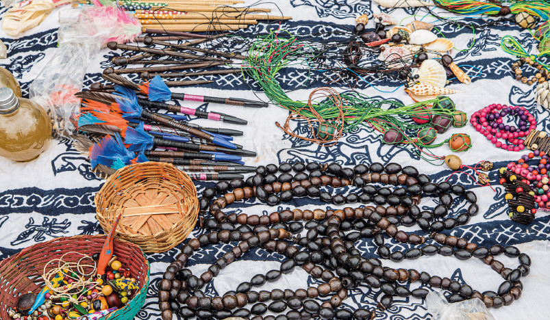 Imagem: Fotografia. Destaque de um tecido estampado estendido e sobre o qual estão vários itens dispostos, como colares de contas, cestos de palha com adereços de contas coloridas, colares de linhas com pedras, pulseiras de contas e palitos de cabelo com penas.  Fim da imagem.