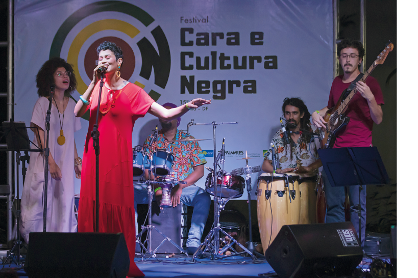 Imagem: Fotografia. No palco, um grupo de músicos se apresenta. A cantora está à frente com um microfone no tripé Ao fundo, uma mulher também canta com instrumentos na mão, um homem de pé toca baixo e dois percussionistas estão sentados. Fim da imagem.