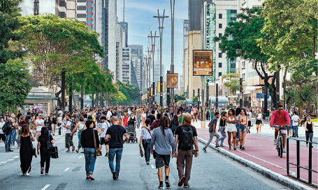 Imagem: Fotografia. Plano aberto de uma ampla via pela qual transitam muitos pedestres. Do lado direito, há uma ciclovia. Ao fundo, árvores, postes e prédios.  Fim da imagem.