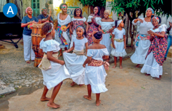 Imagem: A. Fotografia. Em uma área externa, há o destaque de três meninas descalças, de vestido branco com faixa na cintura e lenço no cabelo que dançam em roda. Ao redor delas, outras mulheres com roupa semelhante as observam ao lado de duas ritmistas que tocam atabaques. Fim da imagem.