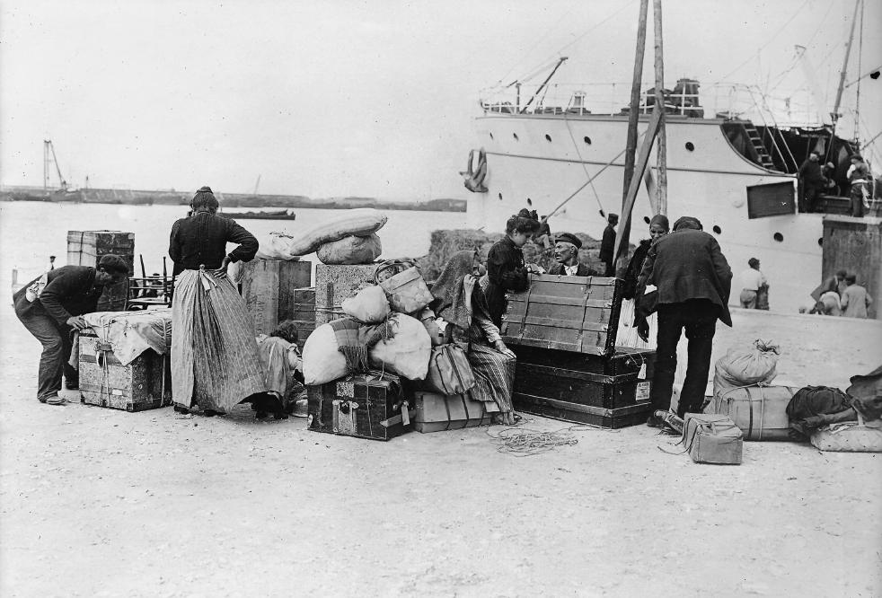 Imagem: Fotografia em preto e branco. Em uma plataforma diante do mar, estão algumas pessoas próximas de muitas malas. Do lado esquerdo, ao lado de baús e trouxas de roupas estão um homem de boina, calça, e paletó que mexe em uma mala, uma mulher de pé, de costas, que usa camisa de manga comprida, saia longa e botas e uma criança.de vestido, blusa de frio, meias e botas. Do lado direito, uma mulher de vestido comprido de manga longa e tecido sobre a cabeça está sentada. Ao seu lado, há um grande baú que uma mulher de longo vestido de manga longa e que está de pé segura aberto. Do outro lado do baú, um homem de paletó, calça e boina ajeita as coisas no chão. Há outro homem e uma mulher próximo. Ao fundo, está a uma grande embarcação próximo da qual estão outras pessoas.  Fim da imagem.