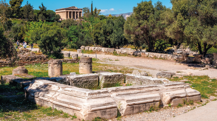 Imagem: Fotografia. Destaque de um local que apresenta chão de terra e grama e ruínas com pedras robustas. Ao redor, há muitas árvores. Fim da imagem.