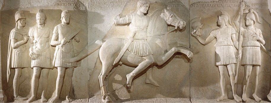 Imagem: Fotografia. Superfície lisa e cinza que apresenta desenhos de homens em relevo. Do lado esquerdo, três homens estão de pé, usam capacete, armadura com saia e possuem espadas. Ao centro, um homem com vestimenta semelhante está a cavalo e tem a espada na cintura. Do lado direito, dois homens com semelhantes estão de pé lado a lado, um deles está com uma das mãos apoiadas em uma lança. Sobre as esculturas há inscrições.  Fim da imagem.