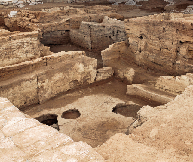 Imagem: Fotografia. Plano aberto de uma paisagem sem vegetação e com ruínas de paredes robustas e altas com crateras no solo. Fim da imagem.