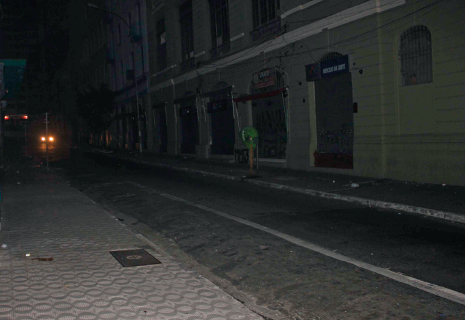 Imagem: Fotografia. Destaque de uma rua e calçada estreitas sem pontos de iluminação. Na via, há um veículo com faróis ligados.  Fim da imagem.