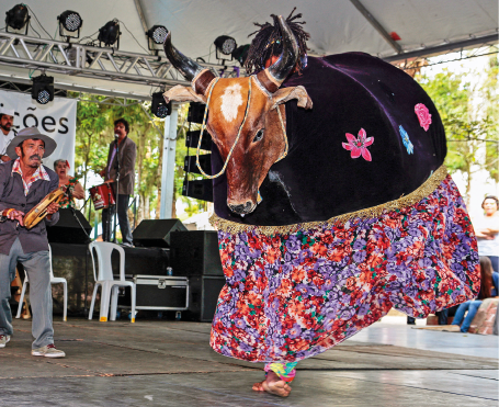 Imagem: Fotografia. Em um palco coberto, destaque de uma pessoa que dança usando a fantasia de um boi preto de cabeça marrom e borda de chita. Ao lado, um rapaz toca um instrumento. Fim da imagem.