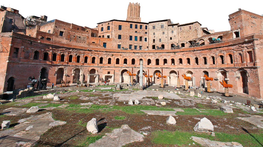 Imagem: Fotografia. Panorâmica do interior de um espaço à céu aberto com chão de pedras e grama e que apresenta uma robusta construção em formato semicircular com arquitetura de arcos na fachada, que são maiores no piso térreo e menores no primeiro piso. Ao centro, há um obelisco. Ao fundo, construções de aspecto similar e que se sobrepõem umas as outras. Fim da imagem.