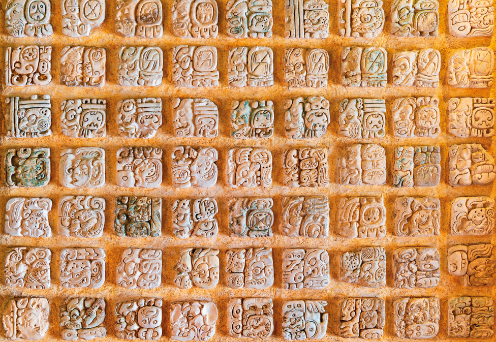 Imagem: Fotografia. Destaque de uma parede com trechos salientes entre linhas com pequenos desenhos em formatos quadrados em relevo. São nove quadros na horizontal e sete quadros na vertical. Fim da imagem.