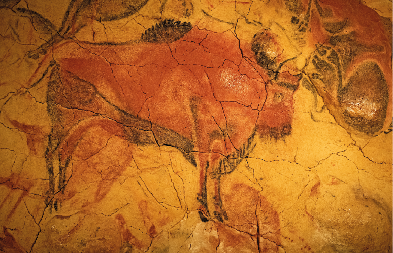 Imagem: Fotografia. Pintura de um animal quadrúpede robusto com chifres em uma parede rochosa amarelada com muitas fissuras.  Fim da imagem.