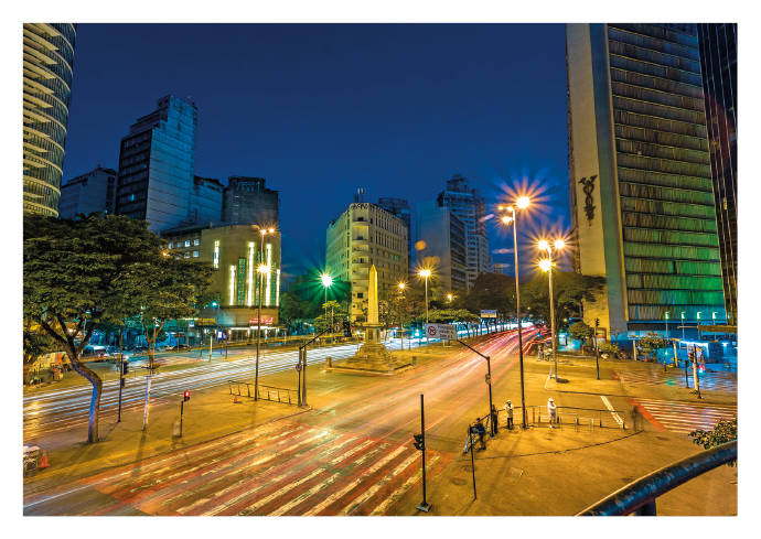 Imagem: Fotografia. Plano aberto de uma ampla avenida bem iluminada durante a noite por meio de postes de iluminação. Ao redor, muitos prédios e algumas árvores. Fim da imagem.