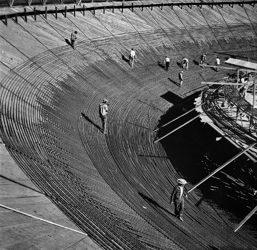 Imagem: Fotografia em preto e branco. Destaque de um local em construção onde estão nove trabalhadores em uma área íngreme e curva com solo com estruturas formando pequenos relevos. Do lado direito, espaço em piso mais alto com vigas e arames.  Fim da imagem.