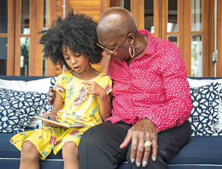 Imagem: Fotografia. Uma senhora de óculos está sentada em um sofá abraçada a uma menina que fala segurando um aparelho eletrônico no colo.  Fim da imagem.