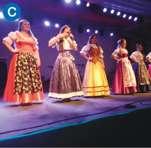 Imagem: C. Fotografia. Sobre o palco, um grupo de mulheres brancas de pé lado a lado com longos vestidos. Elas estão com as mãos na cintura e uma delas segura um microfone.   Fim da imagem.