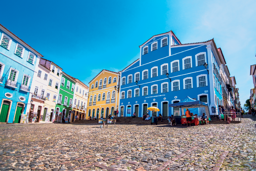Imagem: Fotografia. Um amplo espaço aberto de passagem feito com chão de pedras e rodeado por construções assobradadas com paredes coloridas, amplas janelas e com sacadas. As portas também são coloridas e com acesso diretamente à rua, com calçadas estreitas. Entre as construções, estão estabelecimentos comerciais com produtos expostos do lado de fora e um amplo prédio que apresenta um pequeno conjunto de escadas onde estão sentadas algumas pessoas. Do lado direito, uma pequena barraca de vendas e a continuação da rua ao fundo. Fim da imagem.