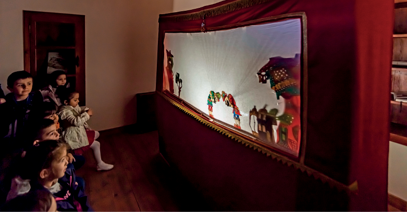 Imagem: Fotografia. Em uma sala escura, muitas crianças sentadas observam um teatro diante de um tecido iluminado. Fim da imagem.