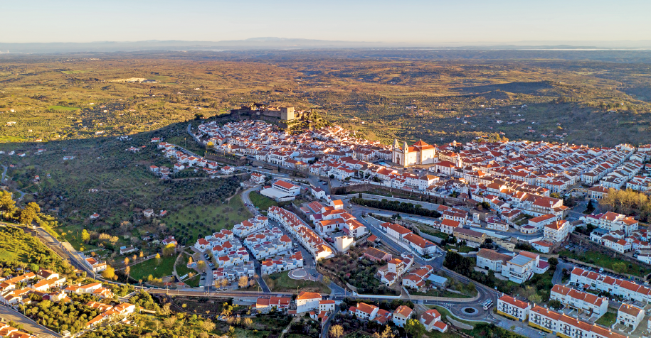 Imagem: Fotografia. Vista aérea de uma pequena cidade rodeada por área verde cultivada e com casas esparsas próximas da cidade. Há uma igreja destacada ao centro e não há construções elevadas.  Fim da imagem.