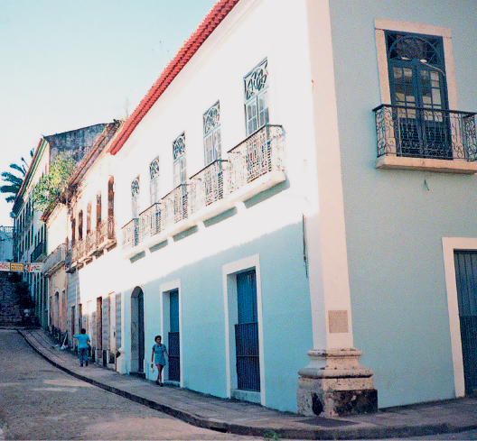 Imagem: Fotografia. Destaque da fachada de uma construção assobradada com janelas com sacadas e portas de acesso diretamente à rua.  Fim da imagem.