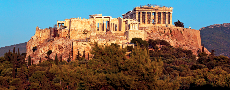 Imagem: Fotografia. Paisagem composta por uma construção robusta em ruínas, sendo que uma parte apresenta muitas colunas. Ao redor, área verde.  Fim da imagem.