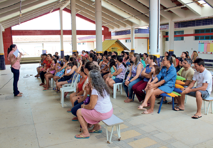 Imagem: Fotografia. Destaque do pátio de uma escola onde homens e mulheres adultos e crianças estão sentados e observam uma mulher falar ao microfone à frente de todos.  Fim da imagem.