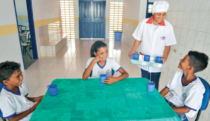 Imagem: Fotografia. Interior de um refeitório onde três alunos uniformizados sentados à mesa sorriem com copos sobre a bancada. De pé, uniformizada e de touca, está uma mulher que também sorri segurando uma bandeja com copos. Fim da imagem.