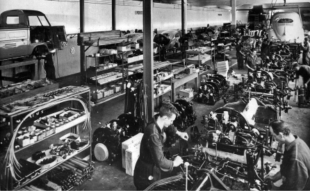 Imagem: Fotografia em preto e branco. Em um amplo galpão, pessoas trabalham com maquinário. No alto, há lataria de carros. Fim da imagem.