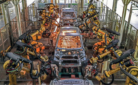 Imagem: Fotografia. Lataria de carros enfileirados sendo soldados por grandes maquinários lado a lado.  Fim da imagem.