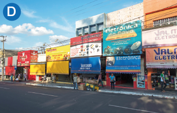 Imagem: D. Fotografia. Destaque de uma rua que apresenta muitos estabelecimentos comerciais com toldos e fachadas com inscrições. No loca, há algumas pessoas.  Fim da imagem.