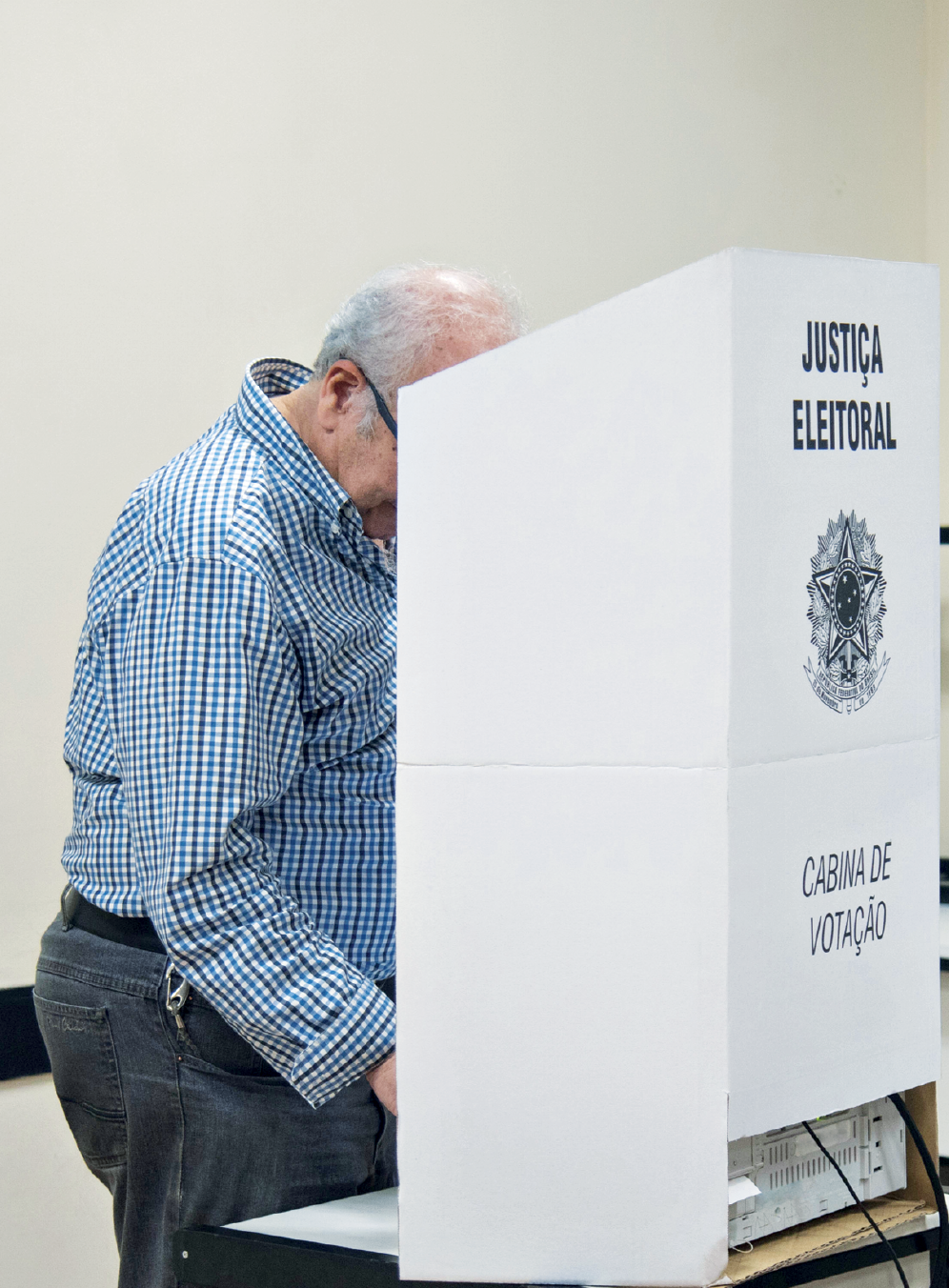 Imagem: Fotografia. Um senhor de pele clara, grisalho, de camisa xadrez e calça está de pé diante de uma mesa que apresenta uma estrutura de uma pequena cabine com a inscrição “JUSTIÇA ELEITORAL. CABINE DE VOTAÇÃO” e um brasão ao centro.  Fim da imagem.