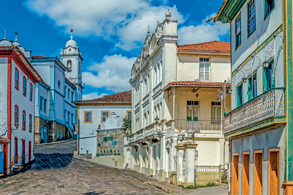Imagem: Fotografia. Destaque de uma rua estreita com paralelepípedos e calçadas bem apertadas com casas assobradadas com batentes coloridos, portas e janelas com acesso diretamente para a via. Algumas construções têm sacadas com parapeito com arabescos e arandelas ao lado das portas. Ao fundo, uma igreja.  Fim da imagem.