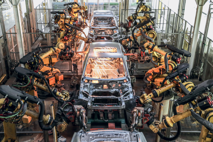 Imagem: Fotografia. Plano aberto de um amplo galpão onde há uma linha de montagem com latarias de automóveis sendo soldadas por grandes máquinas enfileiradas lado a lado. Fim da imagem.