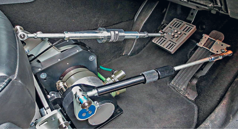 Imagem: Fotografia. Interior de um veículo que apresenta máquinas manipulando os pedais na parte inferior do piso. Fim da imagem.