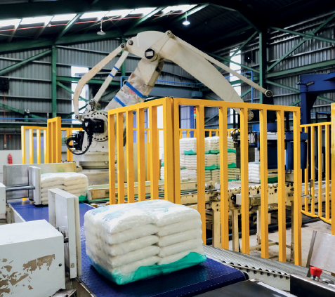 Imagem: Fotografia. Interior de um grande galpão que apresenta uma máquina robótica com longa haste na parte frontal e está rodeado por grandes pacotes, esteiras e divisórias com grades.  Fim da imagem.