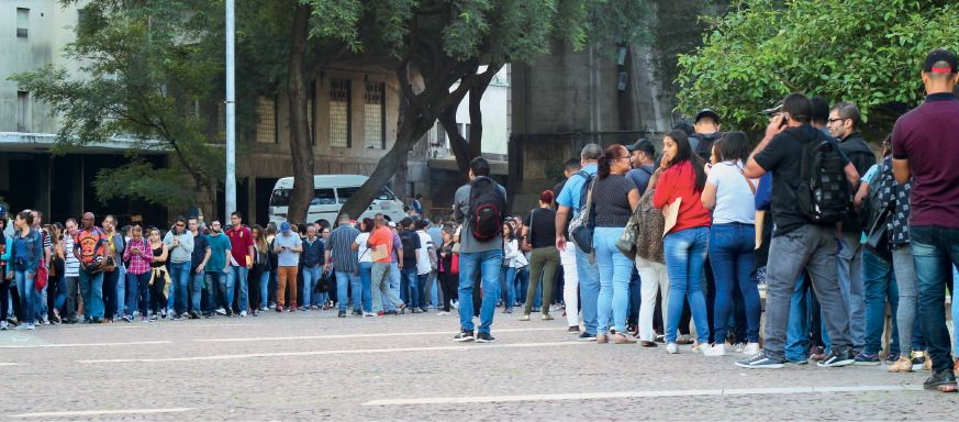 Imagem: Fotografia. Plano aberto de um espaço público onde estão muitas pessoas que formam uma extensa fileira. São homens e mulheres adultos que estão com bolsas e mochilas. Fim da imagem.