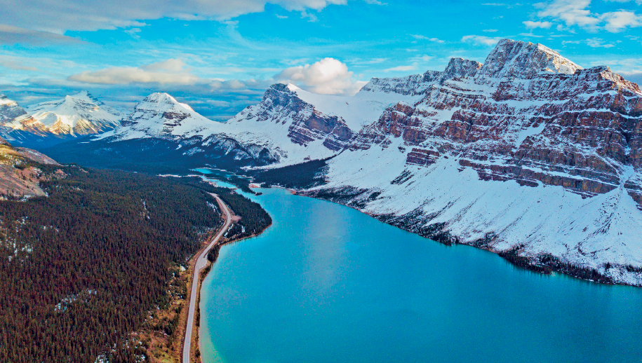 Imagem: Fotografia. Paisagem composta por local com rio caudaloso margeado por mata do lado esquerdo e cadeia montanhosa com neve do lado direito. No horizonte, céu com nuvens.  Fim da imagem.