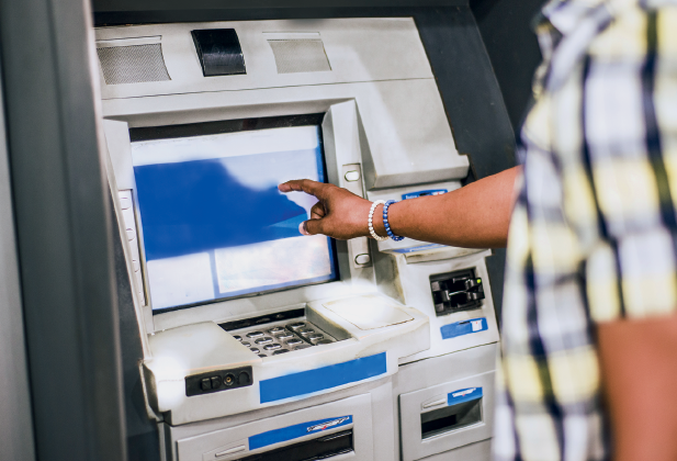 Imagem: Fotografia. Destaque da mão de uma pessoa que está diante de um caixa eletrônico. Fim da imagem.