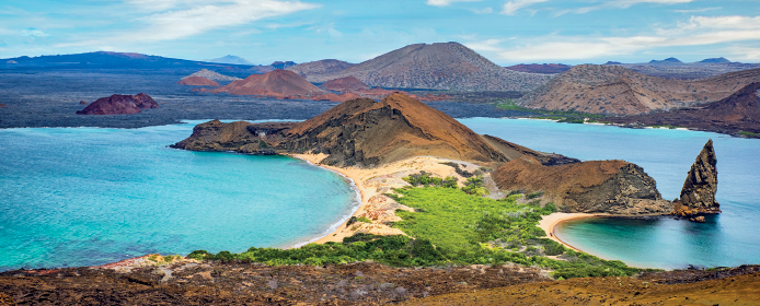 Imagem: Fotografia. Paisagem composta com área peninsular com rochas e área verde margeada por mar límpido e calmo. No horizonte, montanhas e o céu.  Fim da imagem.